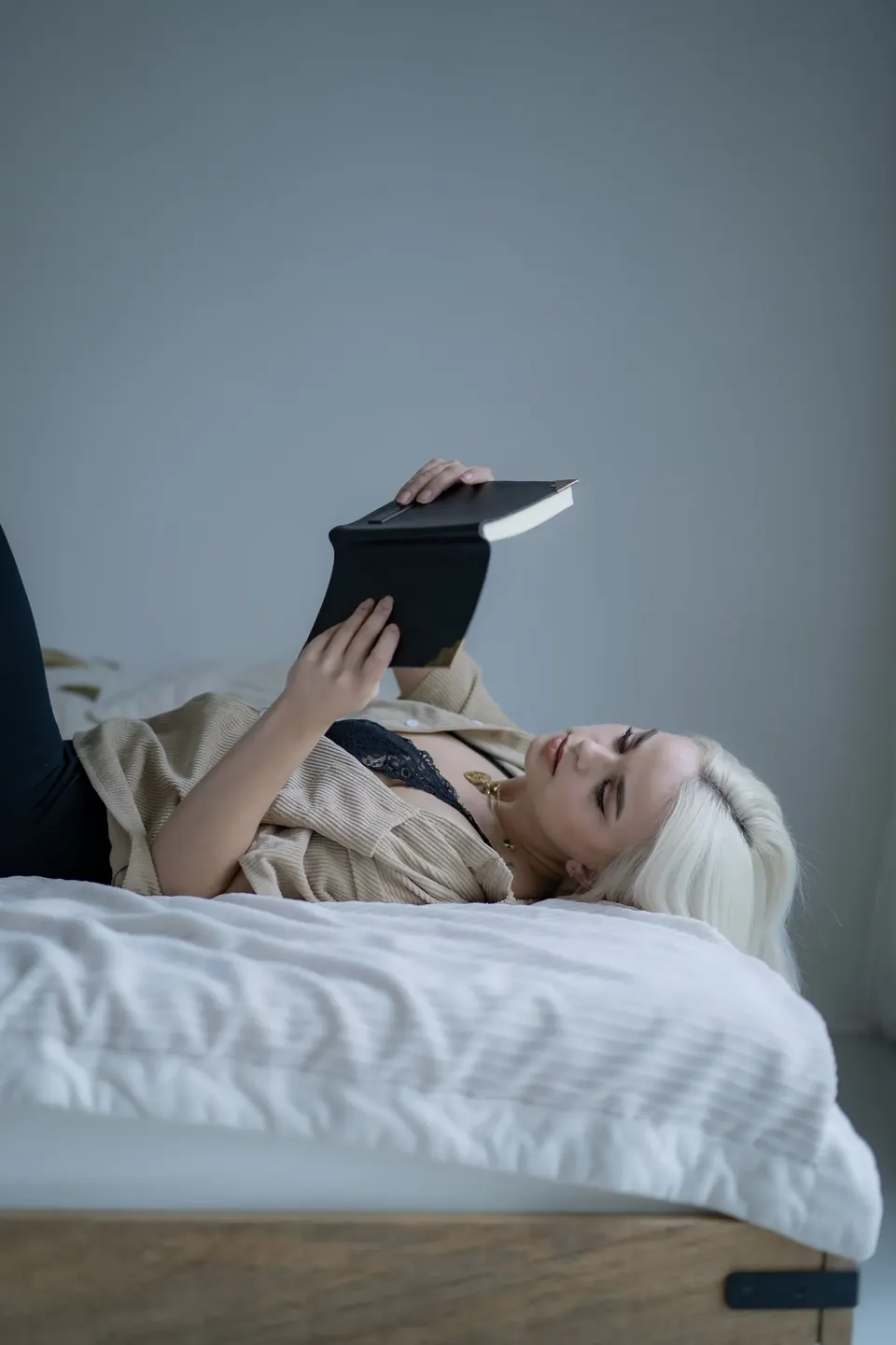 Frau mit platinblonden Haaren liegt auf einem Bett und hält ein Buch über sich