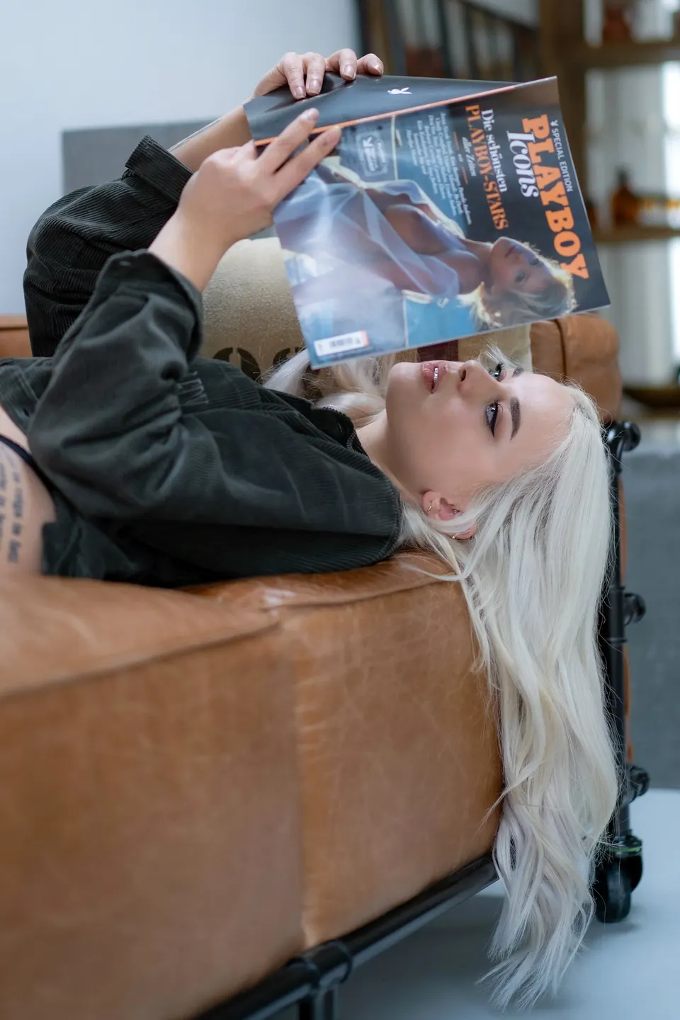 Blonde Frau liegt kopfüber auf einem Ledersofa und liest eine Zeitschrift